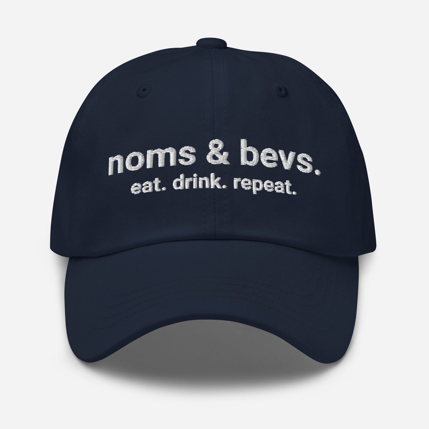 Product mockup of the Navy Blue Hat for Noms & Bevs. On the front of the hat on line one is "noms & bevs." On the second line is "eat. drink. repeat"
