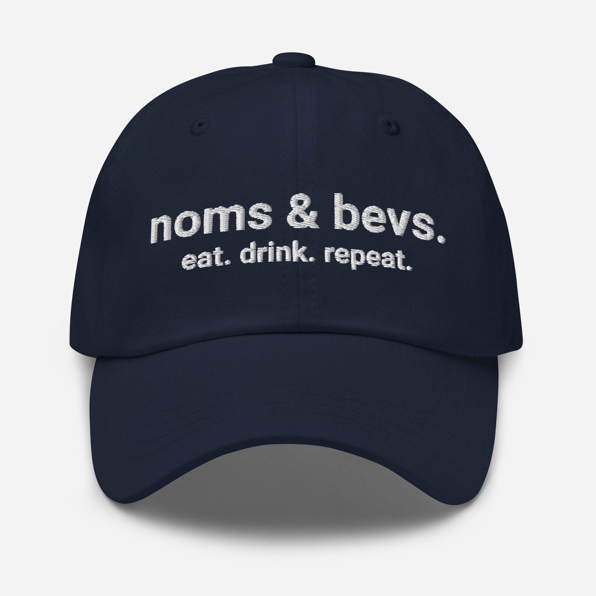 Product mockup of the Navy Blue Hat for Noms & Bevs. On the front of the hat on line one is "noms & bevs." On the second line is "eat. drink. repeat"