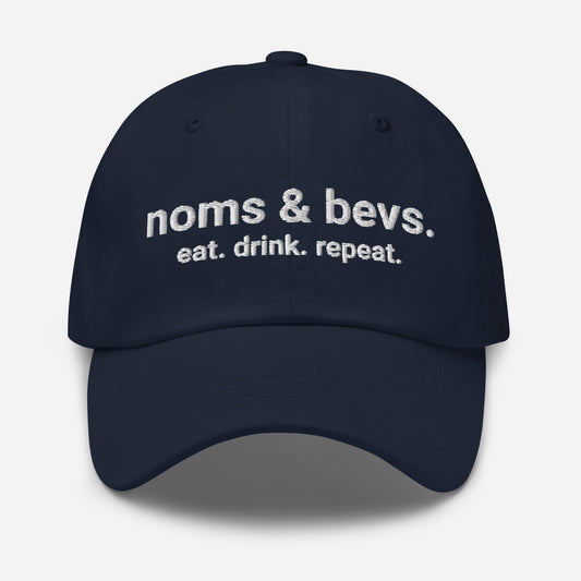 Product mockup of the Navy Blue Hat for Noms & Bevs. On the front of the hat on line one is "noms & bevs." On the second line is "eat. drink. repeat"